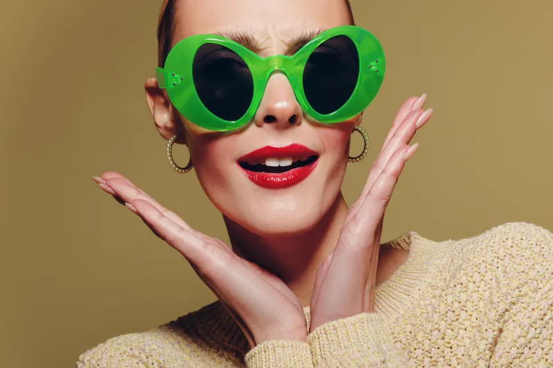 Stylish woman wearing large green sunglasses and red lipstick poses with hands near face. She wears hoop earrings and a beige sweater against a plain background. Fashion and beauty concept.