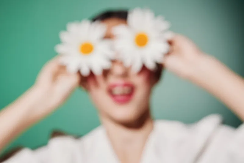 Fashion model holds white daisy flowers over eyes, vibrant natural beauty and creative concept. Soft green background and cheerful face in artistic blur style showcase playful moods.