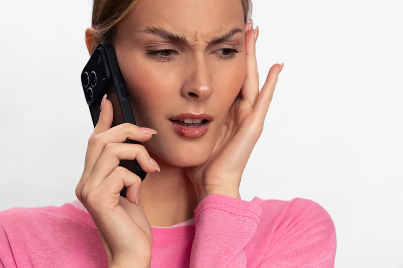 Concerned young woman in pink sweater talks on smartphone, touching face with worried expression. Isolated on white background. Concept for stress, anxiety, communication, and emotion.