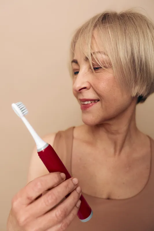 Mature woman smiles while holding red electric toothbrush in beige room. Concept of dental care, oral hygiene, and healthy lifestyle for adults and seniors.