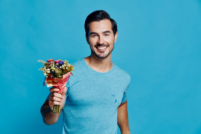 Smiling man holding a colorful bouquet of flowers, wearing a casual blue t-shirt, standing against a bright turquoise background. Concept: joy and celebration.