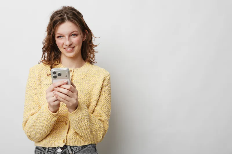 Smiling young woman in yellow sweater holding smartphone, looking away with happy expression. Casual lifestyle, technology use, communication concept on plain light background.
