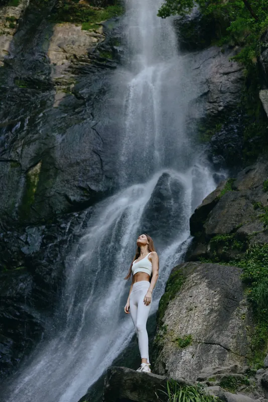 Woman standing near a large waterfall in white fitness wear surrounded by rock formations and lush greenery creating a serene outdoor atmosphere.