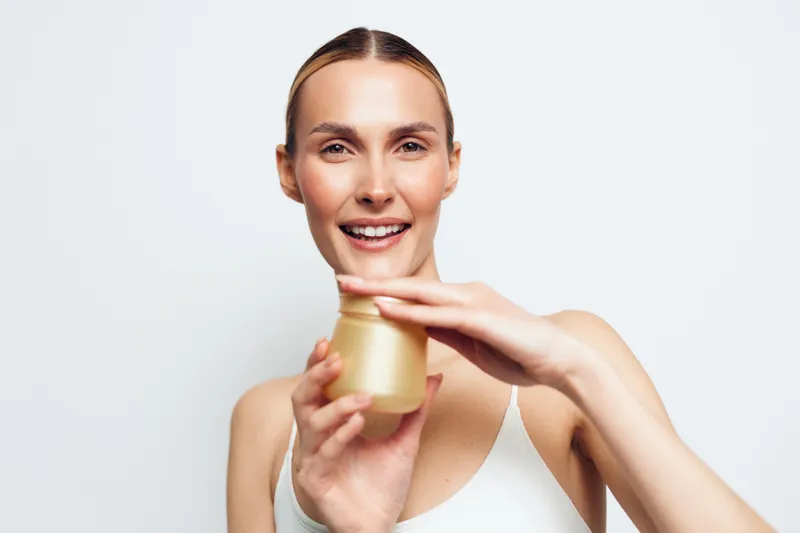 Young woman with pulled back hair smiles while holding a cosmetic jar near her face against a plain white background in a skincare product presentation.