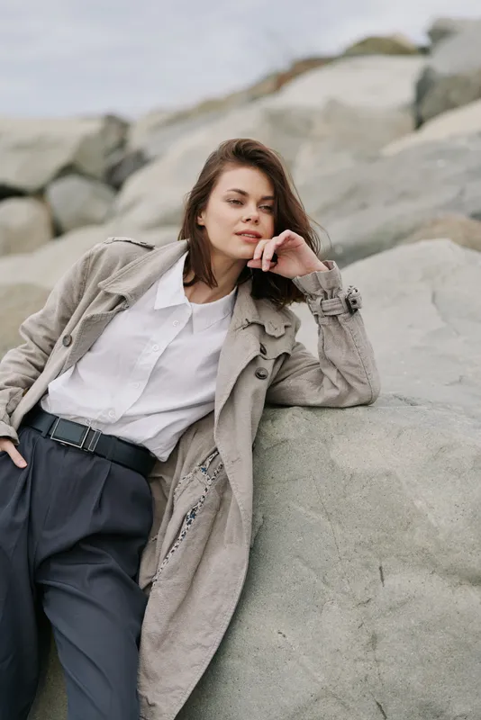 Young woman in stylish coat and white shirt leans on large rocks outdoors. Casual fashion outfit in natural rocky environment with overcast sky. Concept of autumn style and relaxation.
