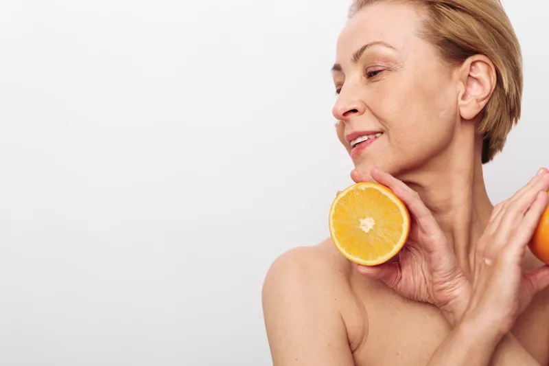 Mature woman with short blonde hair holds fresh orange halves near her shoulder smiling softly on white background. Concept of healthy lifestyle, natural beauty, and wellness care.