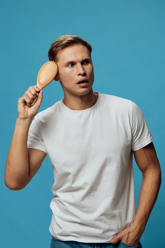 Thoughtful man with surprised facial expression, holding wooden hairbrush near head, wearing casual white t-shirt, standing against vibrant blue background, studio shot, lifestyle concept, contemporar