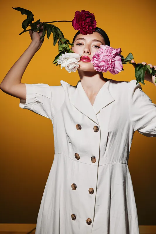 Stylish woman in a white dress holds colorful flowers near her face on a warm orange background. Creative beauty and fashion concept emphasizing elegance and floral accents.