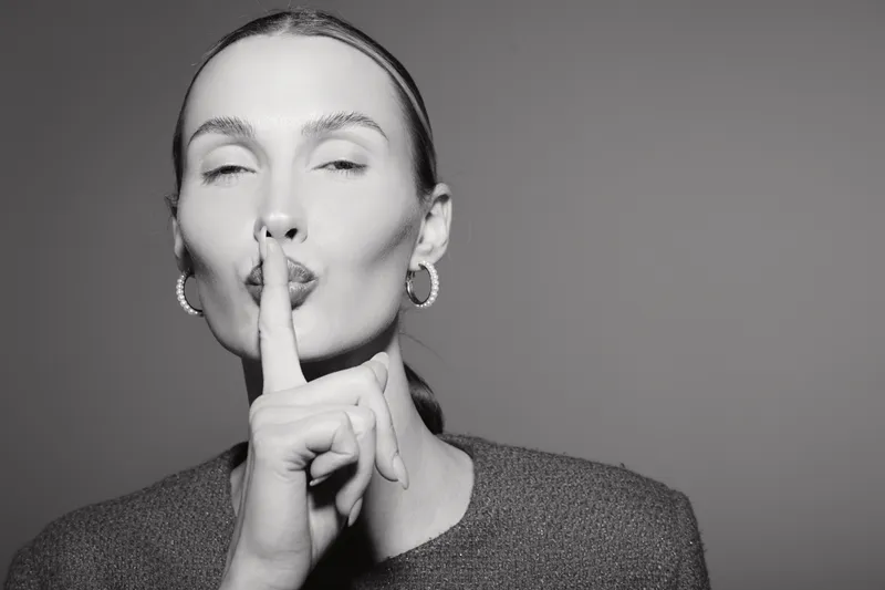 Young white woman with light skin and pulled back hair makes silence gesture with finger on lips. Studio portrait in black and white. Concept of quiet, calm, and secrecy.