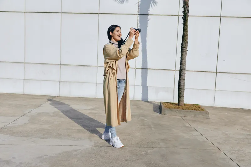 Woman wearing a beige trench coat and jeans takes a photo outdoors with a camera, standing by a pale wall in bright daylight, casual street style and relaxed pose.