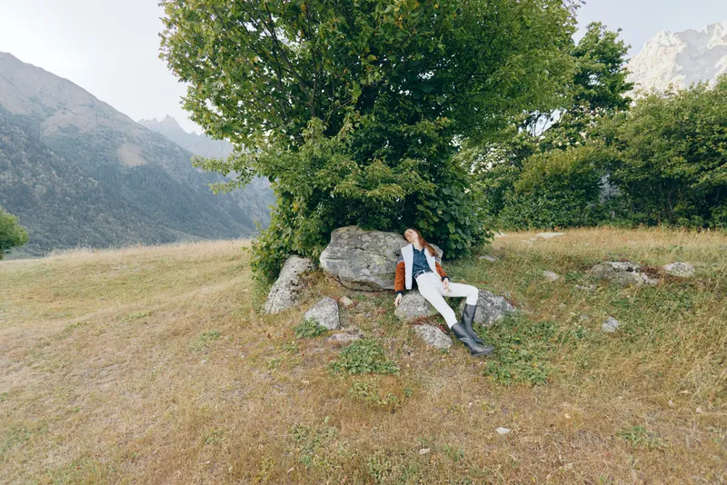 Woman takes a rest on rock under a large tree in a mountain meadow, hiking with backpack and boots, enjoying panoramic mountains view, outdoor relaxation and alpine trek.