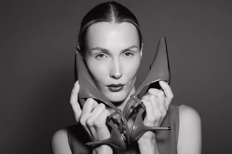 White woman with pale skin holds red high heel shoes near her face in black and white studio portrait. Concept for fashion, elegance, and female style beauty.