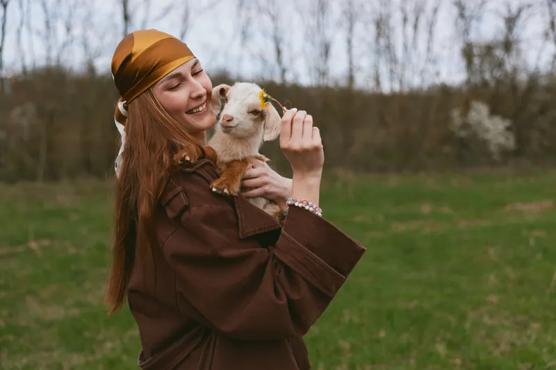 Young woman in a brown coat and headscarf smiles while holding a small goat in a green field during early spring outdoors.