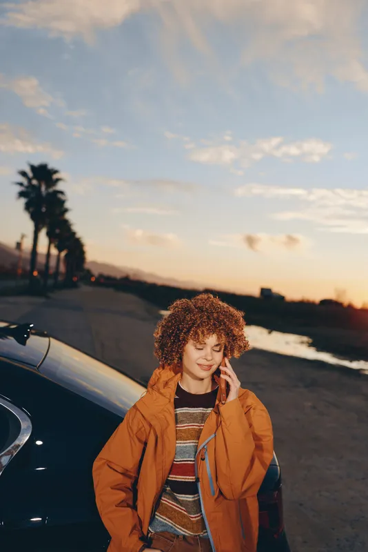 Woman lifestyle portrait at sunset, curly haired woman stands by car near ocean shoreline, orange jacket and striped sweater, coastal road with palm trees, casual travel mood