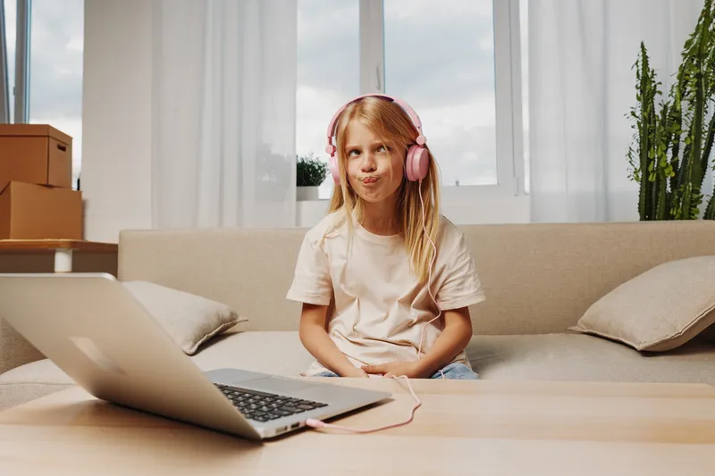 Blonde girl with pink headphones sits on beige sofa, making funny face while using laptop on wooden table in bright living room with plants and cardboard boxes.