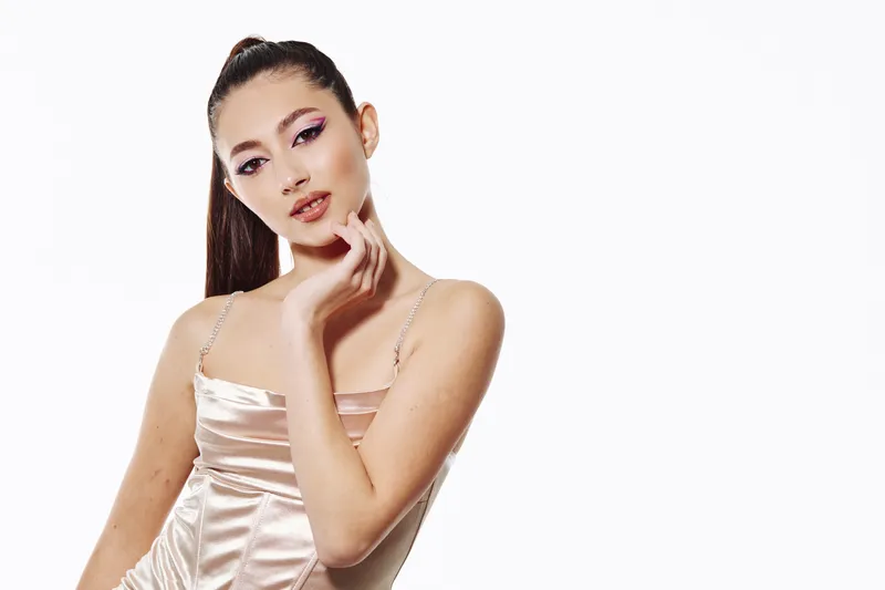 Fashion model with high ponytail and dramatic pink eye makeup poses in soft satin dress against white background. Concept of beauty fashion creative makeup style.