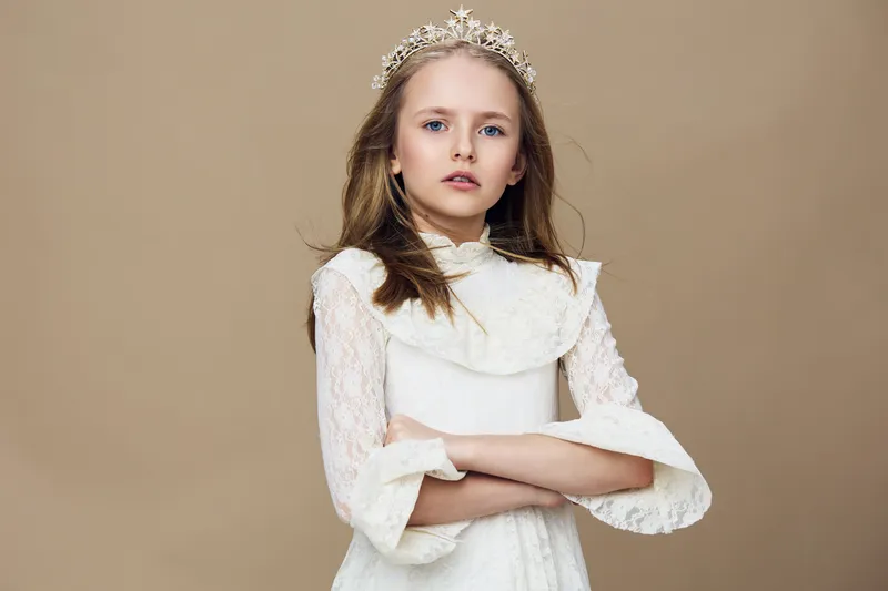 Elegant young girl wearing a lace dress and tiara with a serious expression against a soft beige background, perfect for children s fashion trends and beauty themes