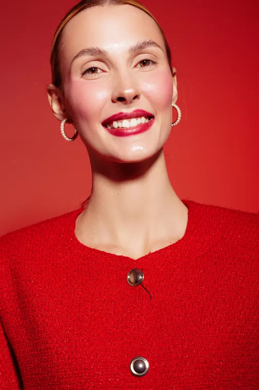 Smiling woman with red lipstick and hoop earrings wears red textured top against red background. Close-up portrait with glossy skin and bright makeup. Concept of beauty and fashion.