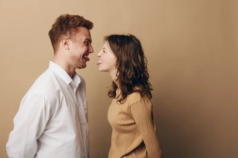 Happy young couple facing each other smiling and laughing in casual clothes against beige background. Concept of love, relationship, joy, and positive emotions between people.
