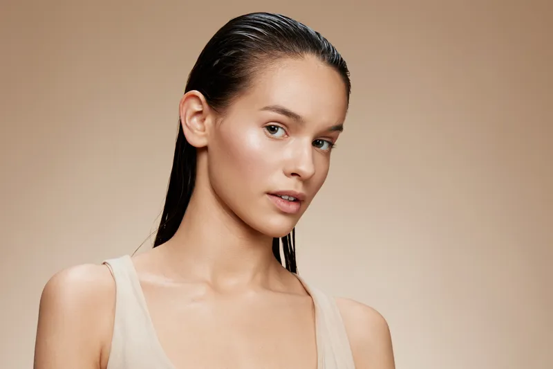 Natural beauty portrait of a young model with wet hair, showcasing glowing skin and subtle makeup against a warm, neutral background