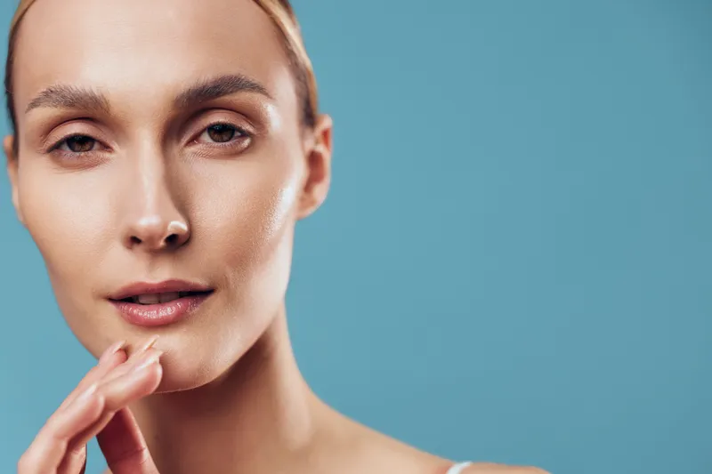 Close-up portrait of a woman with clear skin touching her face gently against a blue background. Concept of skincare, beauty, health, and natural wellness with soft lighting.