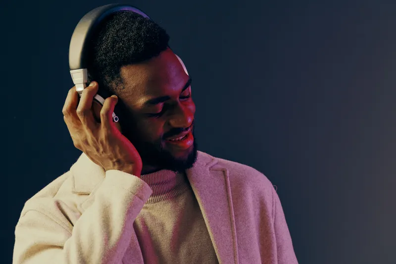 Young Black man enjoying music with headphones, wearing a cozy sweater and stylish coat, showing a joyful expression in a modern indoor setting