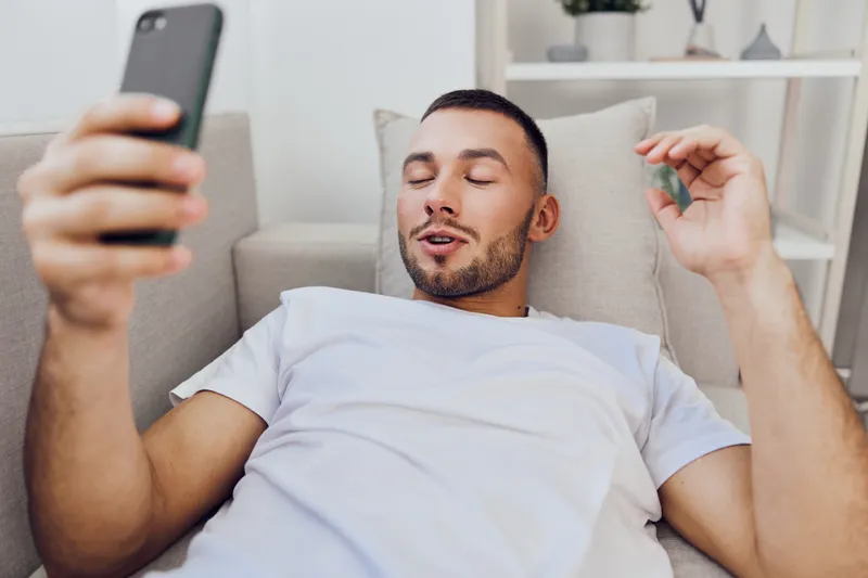 Relaxed man enjoying smartphone technology while lounging on a couch, showcasing modern lifestyle and digital communication trends in contemporary living spaces.