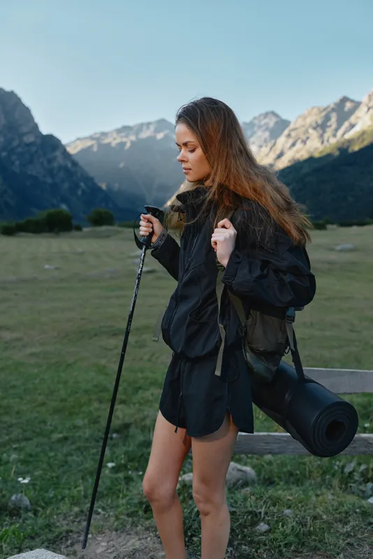 A young woman hikes through a rugged mountain landscape, gripping a trekking pole and carrying a mat. She wears a dark jacket and shorts, focused and calm, enjoying the outdoor adventure.
