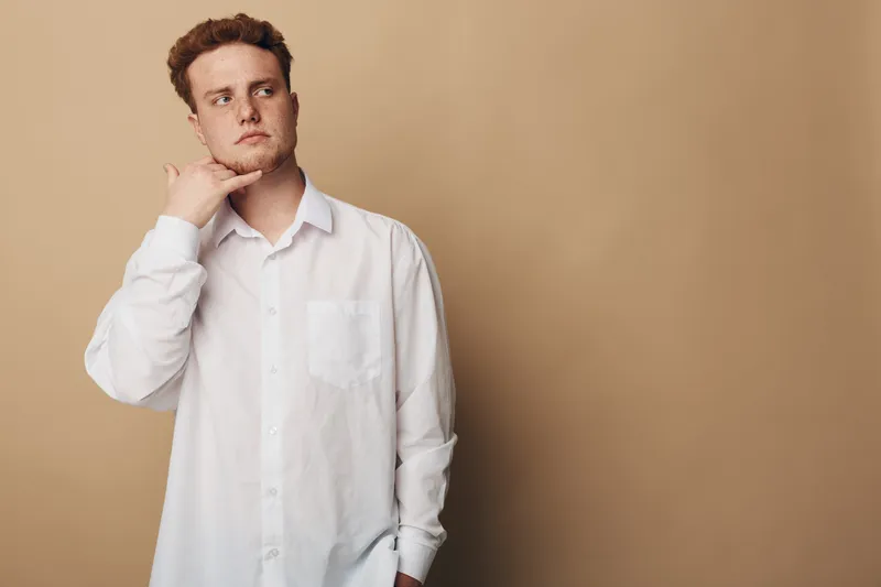 Young man in white shirt makes call me gesture with hand near face, looking away on beige background. Concept of communication, gesture, and casual style.