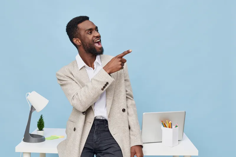 Smiling man with dark skin, curly hair, and a beard pointing to the side, wearing a beige cardigan and white shirt, standing near a modern desk with a lamp, plant, and laptop, isolated on light blue b