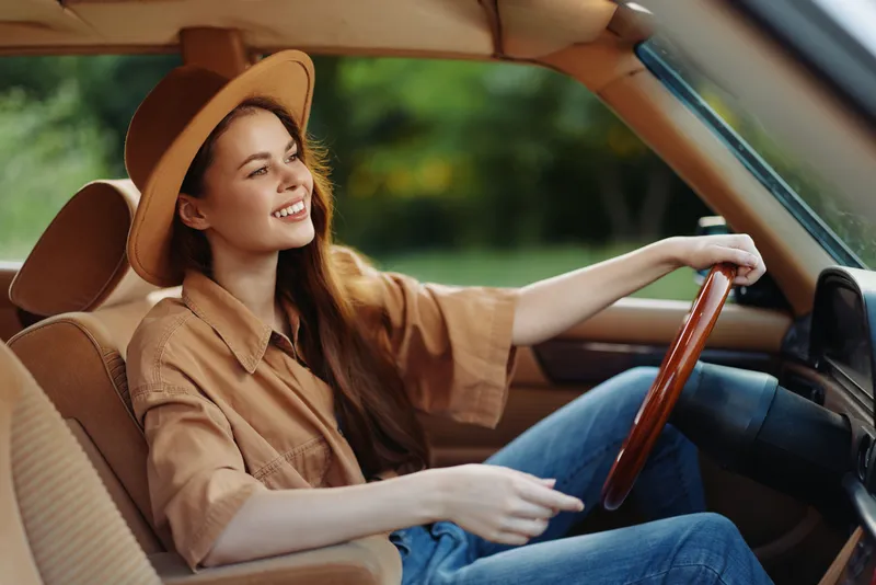 Young woman smiling joyfully in a vintage car, wearing a brown shirt and hat, surrounded by greenery. Perfect for lifestyle, travel and summer themes.
