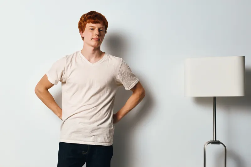 Young man with red hair wearing beige t shirt and dark pants standing confidently indoors near white lamp and wall. Casual style and relaxed posture in minimalistic room setting