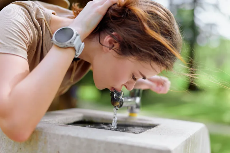 Woman drinking water from a fountain outdoors in a park, casual summer outfit with smartwatch on wrist, closeup of face and hands leaning over stone basin for hydration and refreshment.