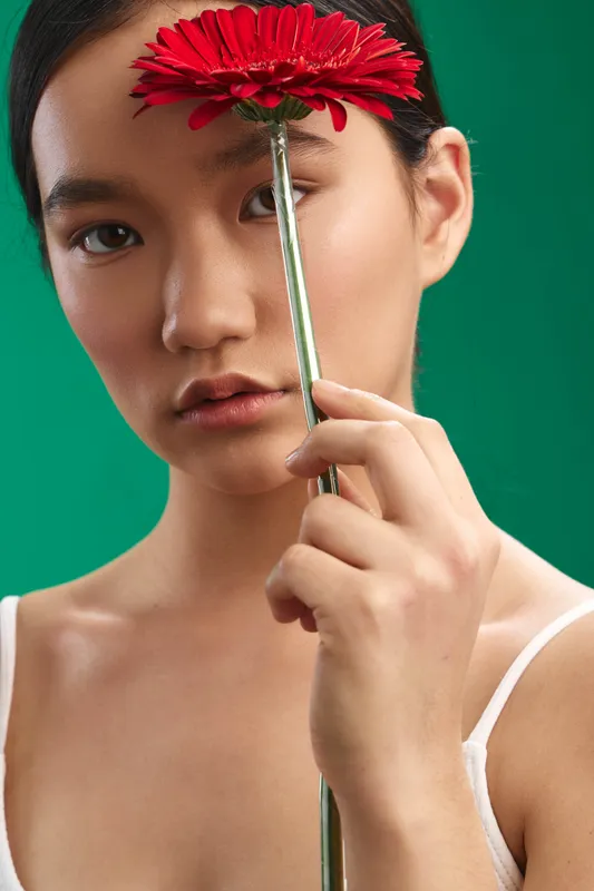 asian woman holds a red flower near her face in a close up portrait with a green backdrop. calm expression and soft lighting create a serene, intimate mood and modern botanical aesthetic.