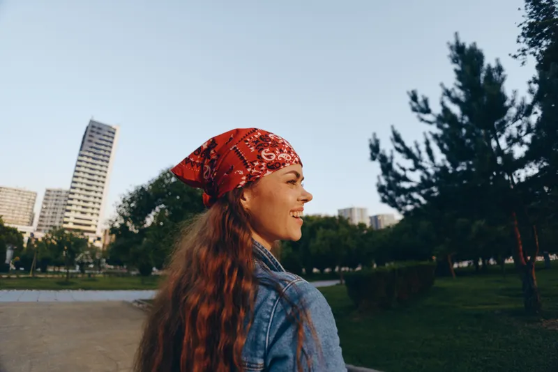 smiling woman with red bandana and long hair wearing denim jacket outdoors in city park with trees and buildings in background during daylight