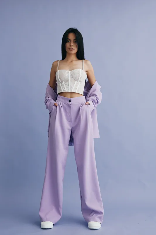 young woman wearing lilac pants and white lace crop top posing confidently against purple backdrop in casual stylish outfit, contemporary fashion look