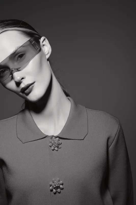 Portrait of a young white woman with pale skin wearing futuristic glasses and a textured sweater with floral buttons. Studio lighting creates dramatic shadows and highlights.
