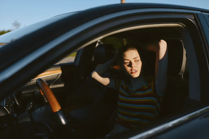 Woman sitting inside a car, striped shirt, thoughtful expression, hands in hair. Close-up on interior. Lifestyle portrait with casual driving mood and retro film color palette. Soft lighting adds
