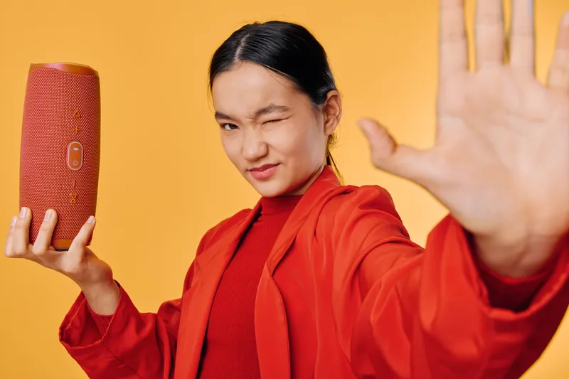 Young woman in red jacket and sweater holding modern wireless speaker while winking with hand raised against yellow background. Portrait of stylish female with technology and casual outfit