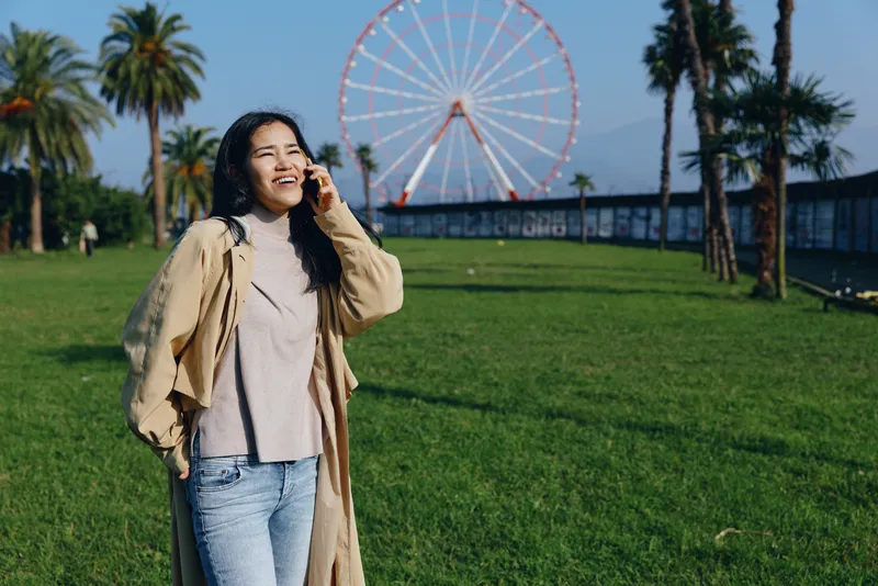 Smiling woman in a light trench coat enjoying a sunny day in a park near a ferris wheel, casual outdoor moment, leisure photography and travel vibe daily.