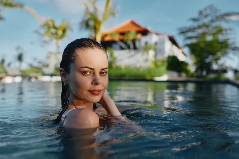 Smiling woman in a pool, sunlit outdoor scene, enjoying a moment of leisure, wellness, and warmth by a resort backdrop.