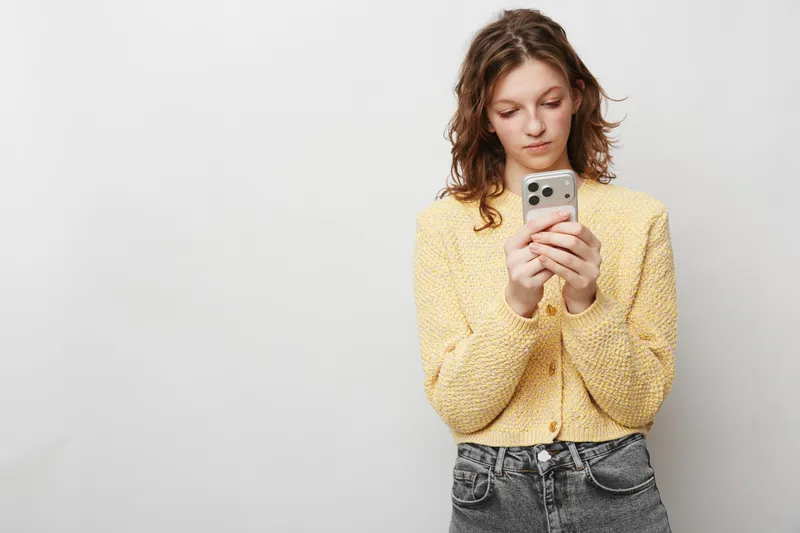 Young woman in yellow sweater looks at smartphone with focused expression, standing against plain white background. Concept of technology, communication, and casual lifestyle.