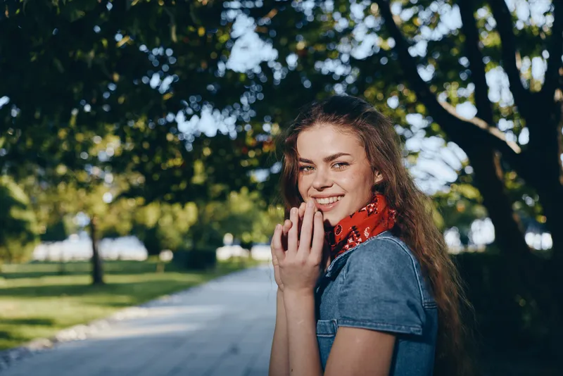 lifestyle woman in modern boho-western denim street style with film color effect, smiling joyfully outdoors under green trees on a sunny day