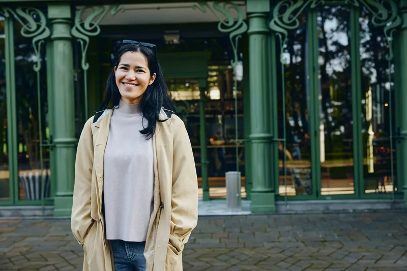Woman in beige coat and gray sweater stands outdoors in front of a green storefront, smiling in an urban daylight scene, blending casual fashion with a relaxed city mood and bright daytime ambiance.