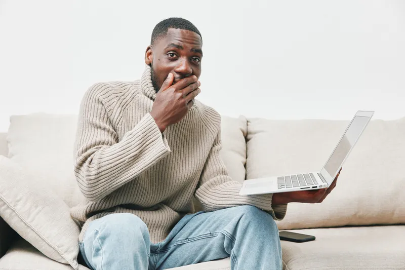 Surprised young black man using a laptop at home, expressing shock and excitement, wearing a cozy sweater, casual indoor setting with neutral colors, ideal for technology concepts