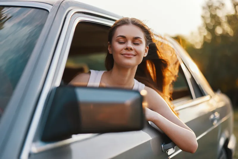 Young woman enjoying a sunny drive with her hair blowing in the wind, reflecting happiness and freedom, captured in a vintage car on a summer day.