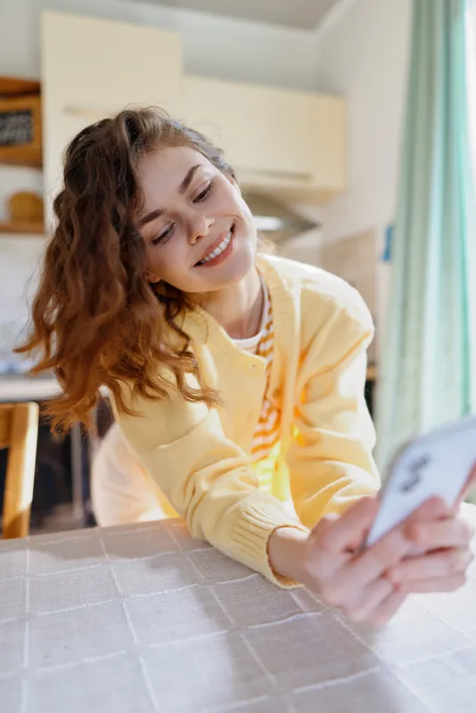 Bright woman indoors, smiling softly while taking a selfie, casual morning moment, sunlit kitchen behind, relaxed pose, warm colors, cozy atmosphere, everyday life at home, natural light mood