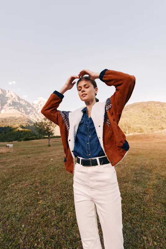 Woman portrait outdoors in mountains meadow, casual jacket and fashion outfit. Young model standing in grassy field with denim shirt and white pants, confident pose in natural landscape.