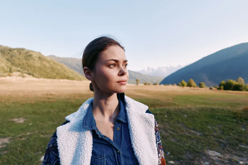 Woman portrait in nature with mountains and meadow, outdoors travel scene. Thoughtful young female stands in wide landscape at golden hour wearing denim and fleece jacket for adventure.
