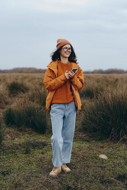 Cheerful woman outdoor lifestyle walking along a grassy path in an autumn field, wearing orange knit jacket and beanie, smiling in soft daylight, relaxed posture, natural tones today gently